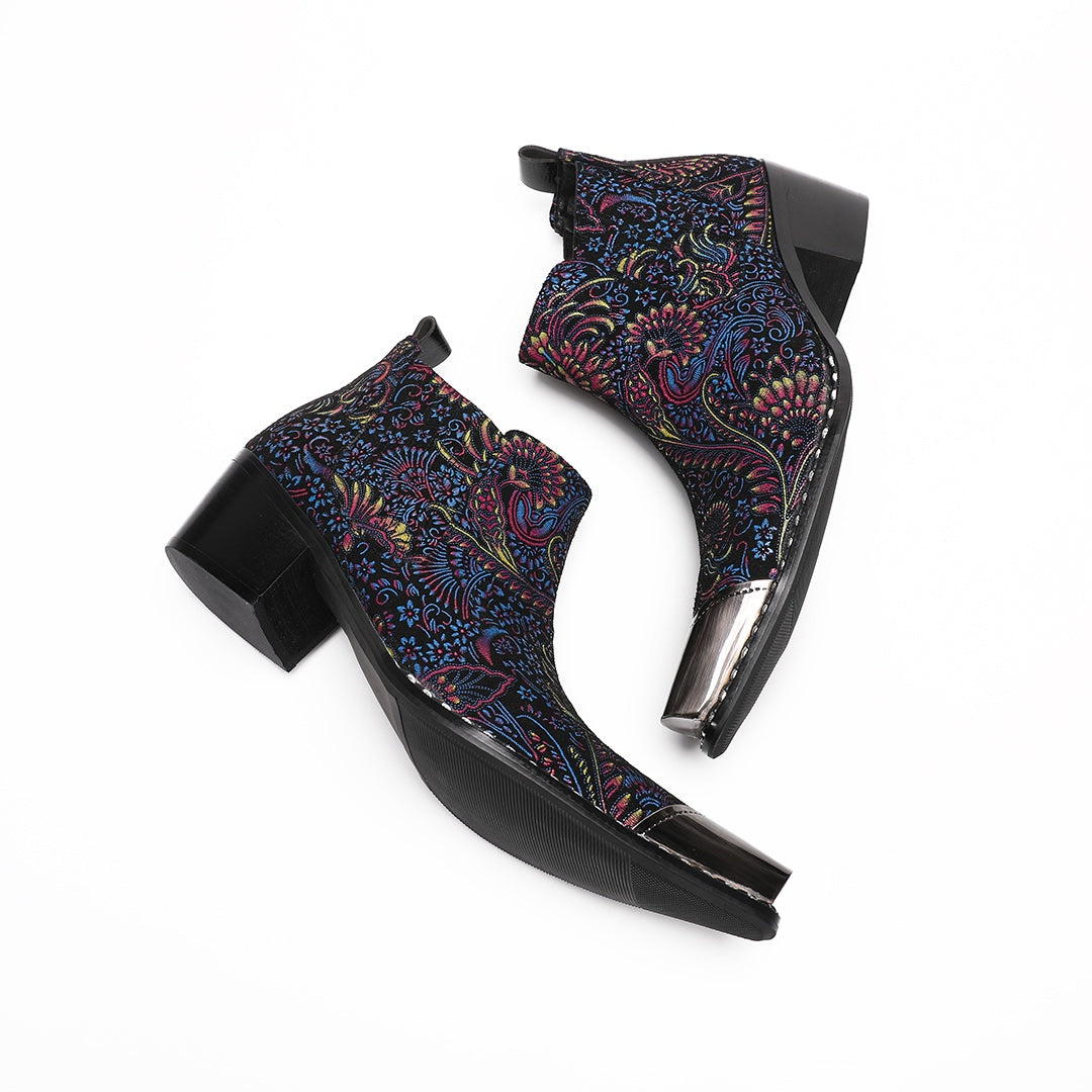 A pair of men's black ankle boots with a vibrant multicolor brocade pattern and a polished silver metal toe cap.